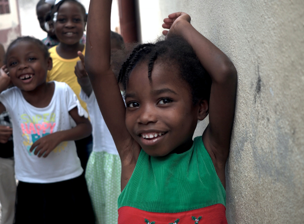 Girl smiles at the camera with her arms raised while other children smile behind her